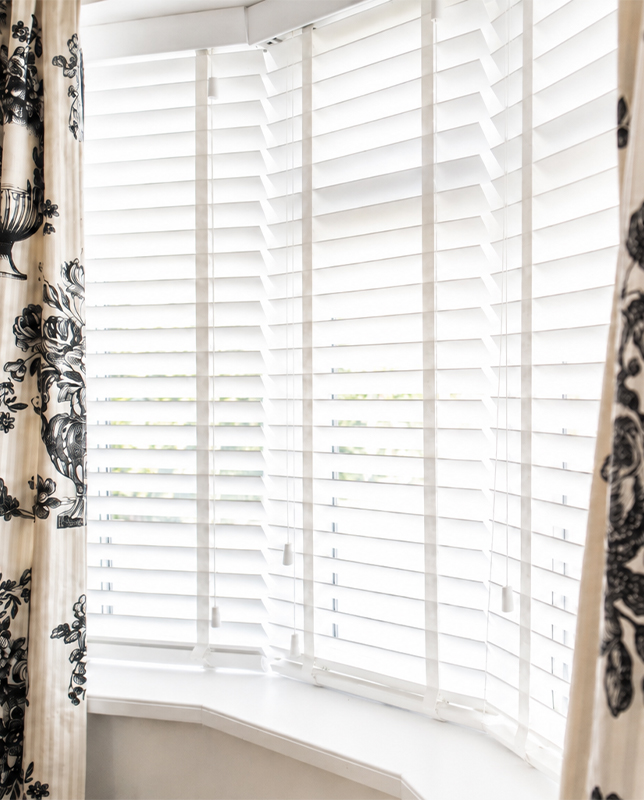 Decorquip white faux wood Venetian blinds fitted to a bay window with patterned curtains in a living room